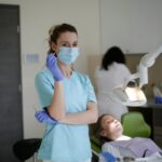 Female assistant and orthodontist in sterile mask and latex gloves taking dental mirror and tweezers while standing near lying female patient and lamp and looking at camera